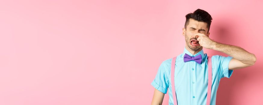 Image of man crying from disgusting smell, shut his nose from awful stink, standing in bow-tie and suspenders on pink background