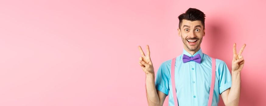 Cheerful young guy with moustache and bow-tie showing peace or victory gestures, smiling happy at camera, standing on pink background