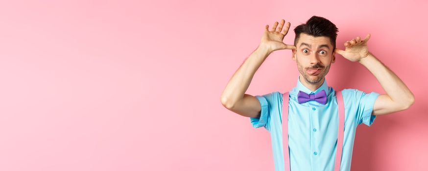 Silly and childish guy in suspenders and bow-tie, mocking someone, fooling around and making funny gestures, standing on pink background