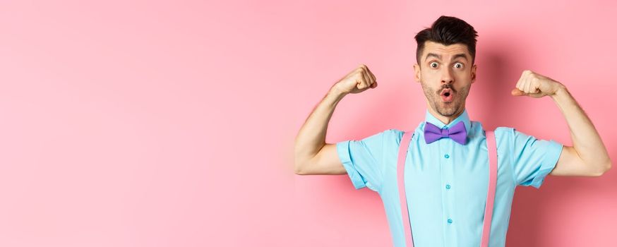 Image of funny guy with moustache and bow-tie, showing muscles, flexing biceps and looking surprised at camera, standing in disbelief on pink background