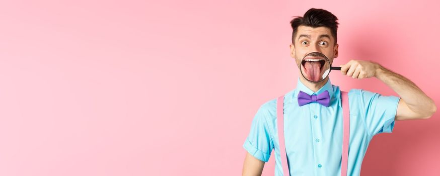 Funny caucasian guy showing tongue in magnifying glass, smiling and making faces, standing in bow-tie over pink background
