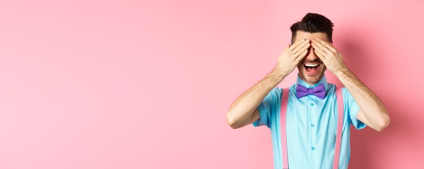 Cheerful young man celebrating, waiting for surprise with closed eyes and happy smile, standing joyful on pink background