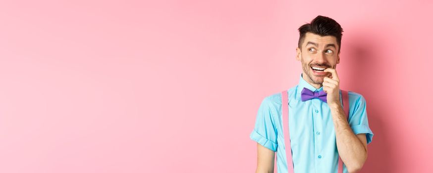 Image of smiling man making choice, looking dreamy and happy aside, biting fingernail with tempted expression, wanting something, standing over pink background