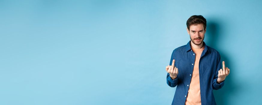 Ignorant and unbothered man showing middle-fingers and looking reluctant at camera, give zero fucks, standing on blue background