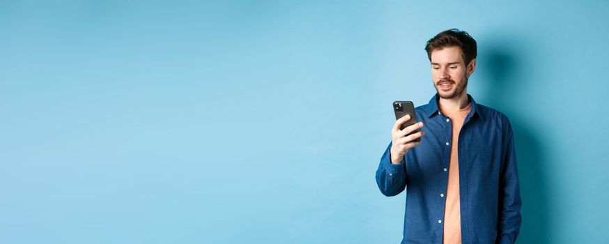 Handsome modern guy using mobile phone, reading smartphone screen and smiling, networking on blue background