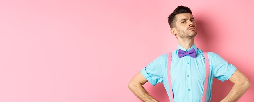 Proud and confident caucasian guy with moustache and bow-tie, looking arrogant with chin up, frowning and looking at camera, standing on pink background