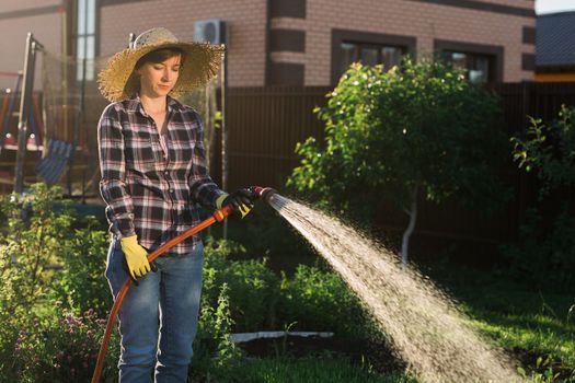 Caucasian woman gardener in work clothes watering the beds in her vegetable garden on sunny warm summer day. Concept of working in the garden and your farm