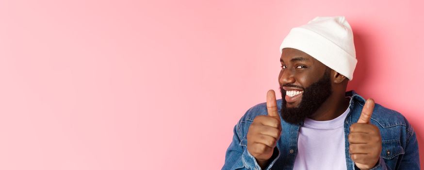 Close-up of happy Black bearded guy in beanie showing support, agree or approve something, giggle devious and showing thumbs-up, standing over pink background