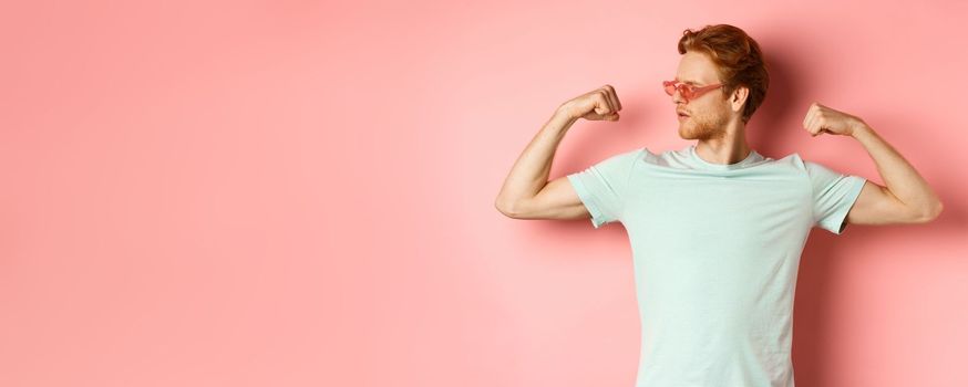 Confident young man with red hair, wearing summer sunglasses and t-shirt, showing strong and fit body muscles, flex biceps and staring cool at camera, pink background