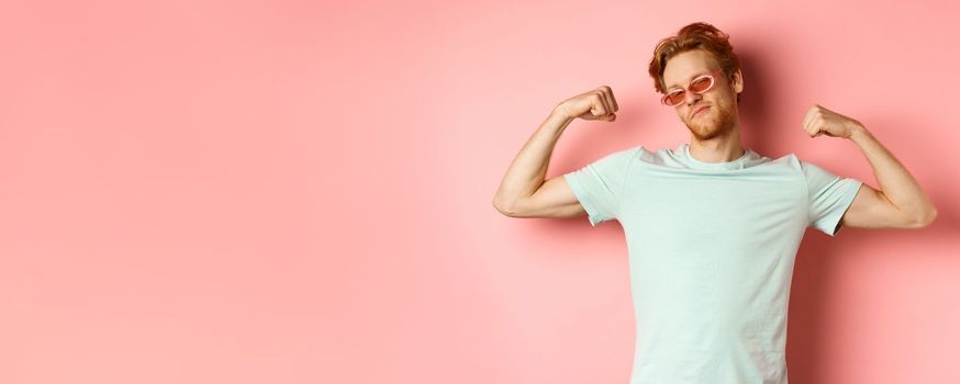Confident young man with red hair, wearing summer sunglasses and t-shirt, showing strong and fit body muscles, flex biceps and staring cool at camera, pink background