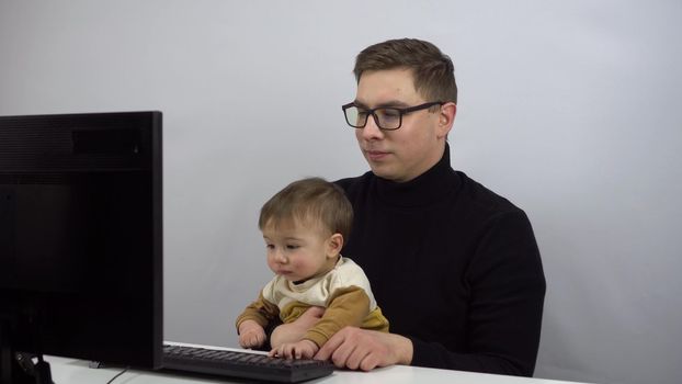 The IT specialist works remotely with his son in his arms. A young man in glasses with a one-year-old son is typing on a pc.
