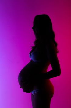 Beautiful young pregnant woman in lingerie is happily stroking her belly in studio in beautiful neon purple lighting. Concept of beauty and expectation of meeting with your beloved baby