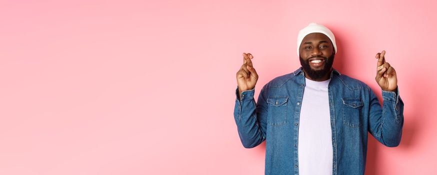 Optimistic african-american man making wish, holding fingers crossed and smiling, standing over pink background