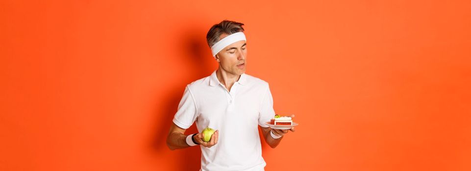 Portrait of middle-aged sportsman making decision between green apple and cake, being on diet, standing over orange background