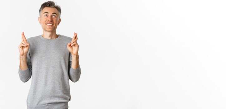 Image of excited and hopeful middle-aged man making a wish, begging God, cross fingers for good luck and looking up, standing over white background
