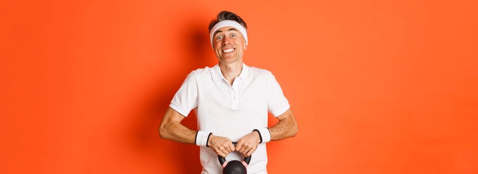 Concept of workout, gym and lifestyle. Portrait of funny middle-aged sportsman trying to lift kettlebell, standing over orange background
