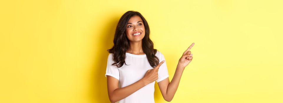 Portrait of beautiful african-american woman, smiling and looking dreamy at promo, pointing fingers at upper left corner, showing logo over yellow background