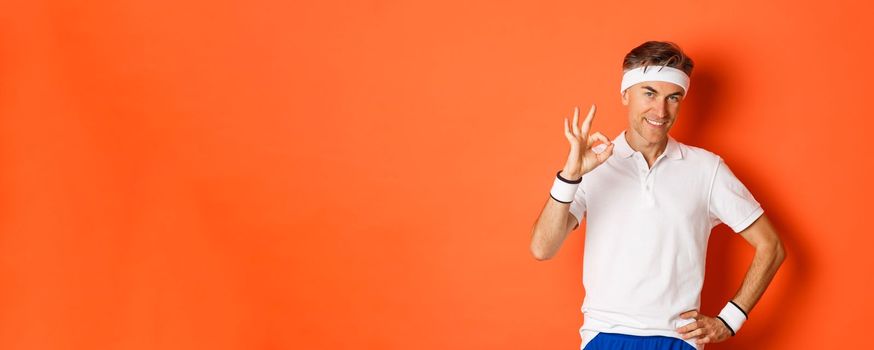 Concept of sport, fitness and lifestyle. Portrait of confident middle-aged sportsman, showing okay sign and look pleased, guarantee something or recommending gym, orange background