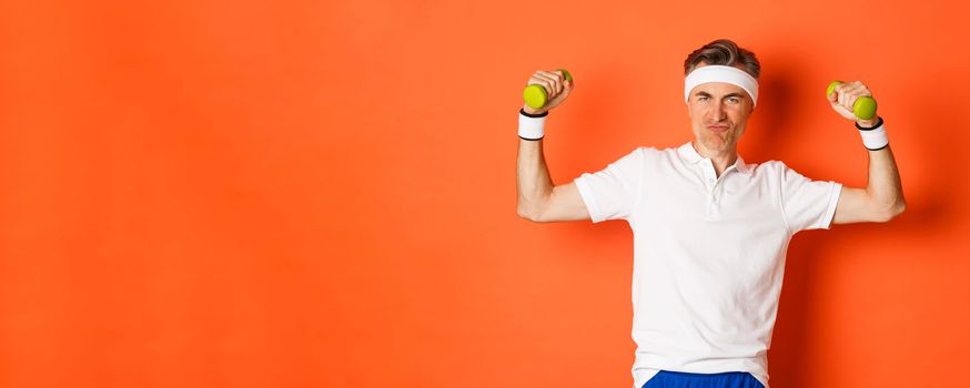 Portrait of cheeky middle-aged fitness guy, doing sports against orange background, flex biceps and holding dumbbells