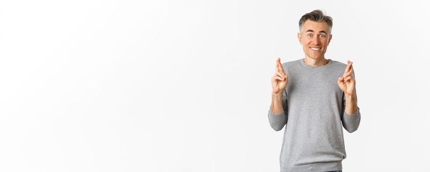 Image of excited and hopeful middle-aged man making a wish, waiting for results, cross fingers for good luck and smiling, standing over white background