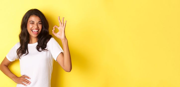 Portrait of confident and attractive african-american girl, have everything under control, showing okay sign and winking to assure you, approve something good, posing over yellow background