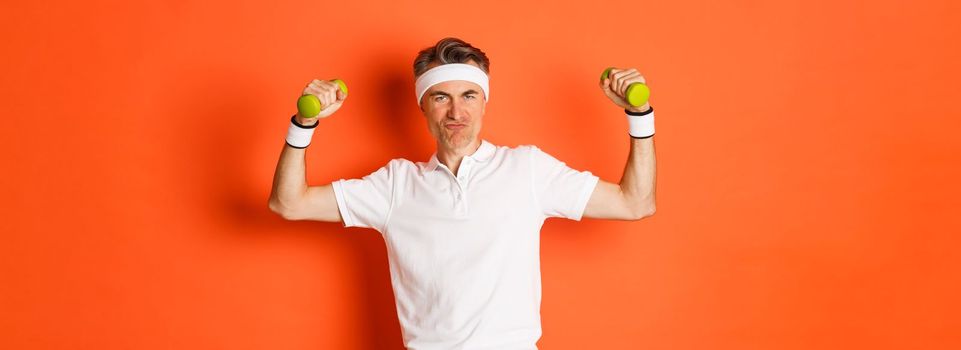 Portrait of cheeky middle-aged fitness guy, doing sports against orange background, flex biceps and holding dumbbells