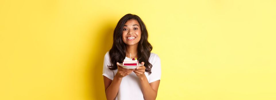 Portrait of beautiful african-american girl celebrating birthday, smiling and looking happy and holding b-day cake with candle, standing over yellow background