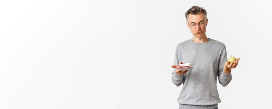 Portrait of thoughtful middle-aged man in glasses, making decision between tasty cake and green apple, standing over white background