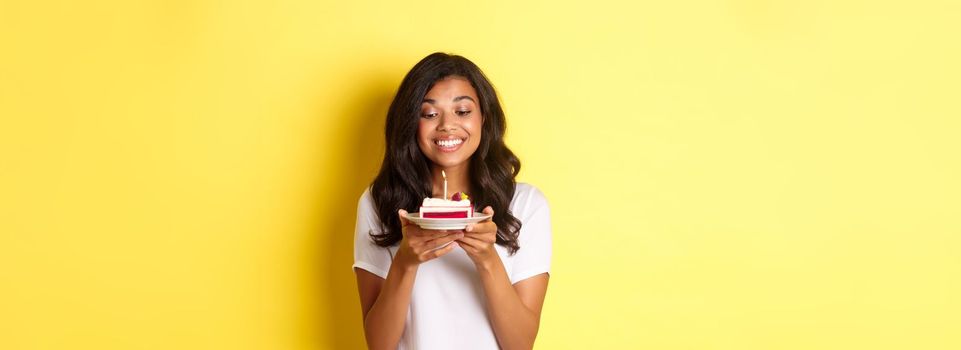 Portrait of beautiful african-american girl celebrating birthday, smiling and looking happy at b-day cake with candle, standing over yellow background