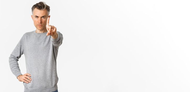 Image of serious-looking and confident middle-aged man telling no, showing stop or taboo gesture, extend one finger to prohibit something, standing over white background