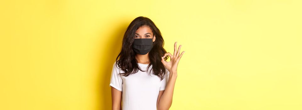 Concept of coronavirus, pandemic and lifestyle. Portrait of smiling african-american woman in face mask, showing okay sign in approval, recommend or guarantee something, yellow background