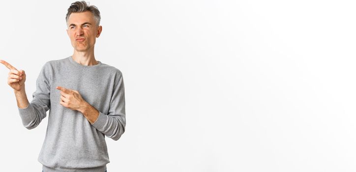 Portrait of skeptical handsome adult man, wearing grey casual sweater, grimacing as looking and pointing at something bad, express negative opinion, standing over white background