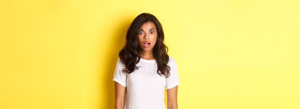 Portrait of shocked and speechless african-american girl, drop jaw and looking at amazing promo offer, standing over yellow background with copy space