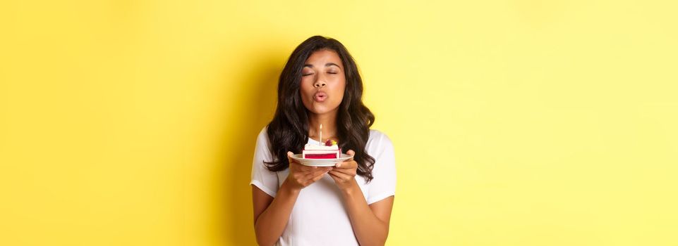 Portrait of beautiful african-american girl celebrating birthday, blowing lit candle on b-day cake and making a wish, standing over yellow background