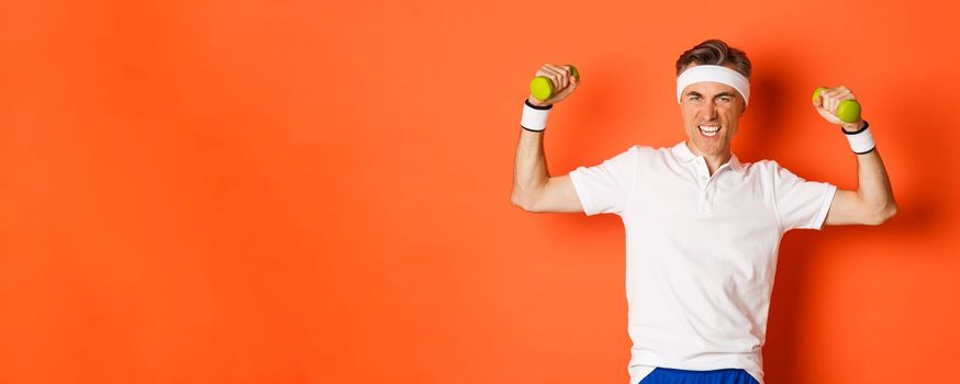 Portrait of confident middle-aged fitness guy, doing sports over orange background, flex biceps and holding dumbbells, showing muscles