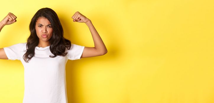 Image of strong and confident african-american girl, flexing biceps and looking self-assured, standing over yellow background
