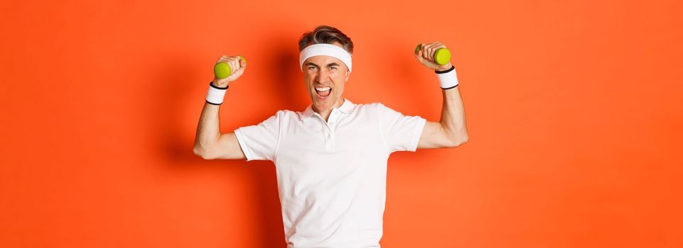 Portrait of confident middle-aged fitness guy, doing sports over orange background, flex biceps and holding dumbbells