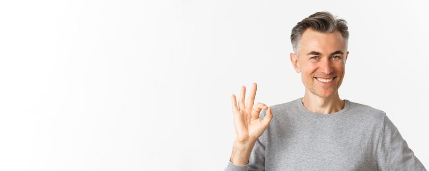Close-up of handsome middle-aged man, showing okay sign and smiling, guarantee quality, assure that everything good, standing over white background