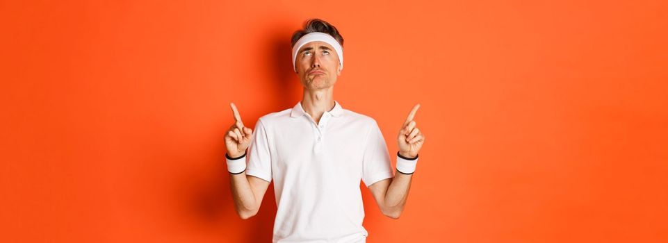 Image of upset and gloomy middle-aged guy, pointing and looking up disappointed, workout over orange background
