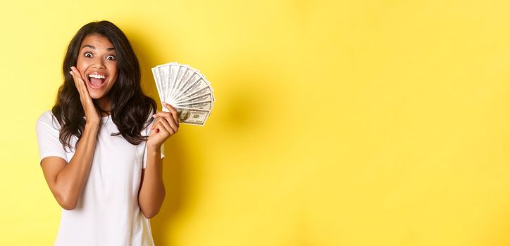 Image of excited lucky girl, winning money and smiling amazed, standing over yellow background