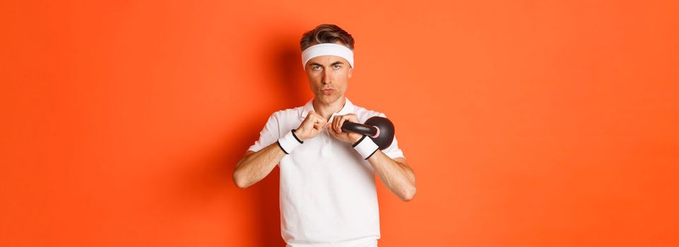 Concept of workout, gym and lifestyle. Close-up of determined middle-aged fitness guy, doing sport exercises with kettlebell, standing over orange background.