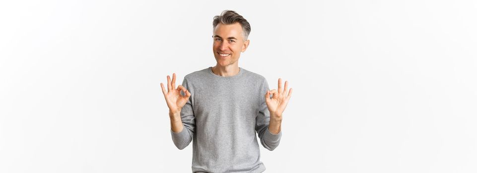 Portrait of handsome middle-aged man, smiling and looking confident while showing okay signs, guarantee something is good, standing over white background