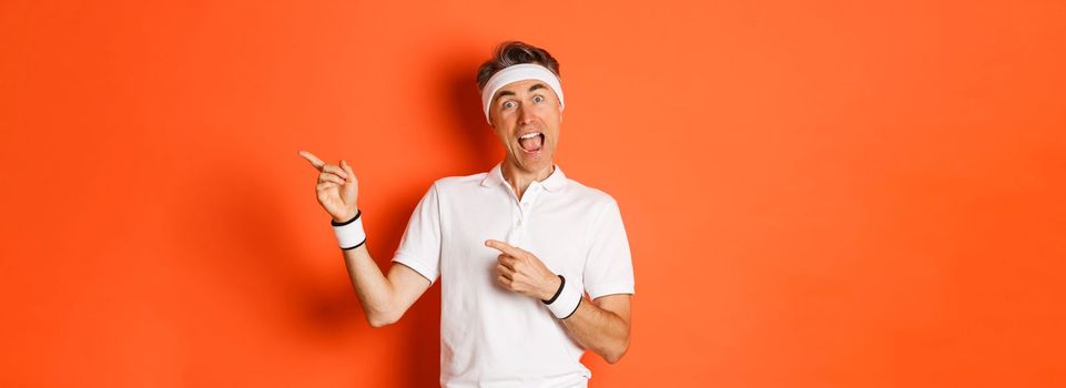 Concept of workout, sports and lifestyle. Image of excited middle-aged male athlete, wearing activewear for exercises, pointing fingers at upper right corner, showing logo, orange background