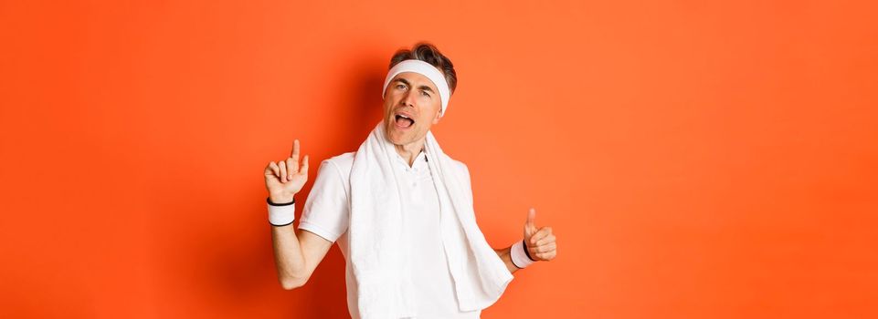 Image of happy and carefree middle-aged fitness guy, dancing after workout, holding towel over neck, posing against orange background.
