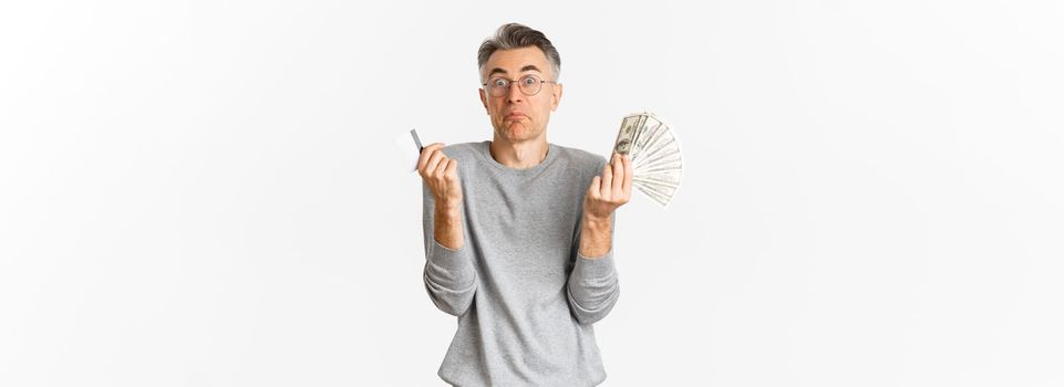 Portrait of clueless middle-aged man in glasses, shrugging while holding credit card with money, standing indecisive over white background