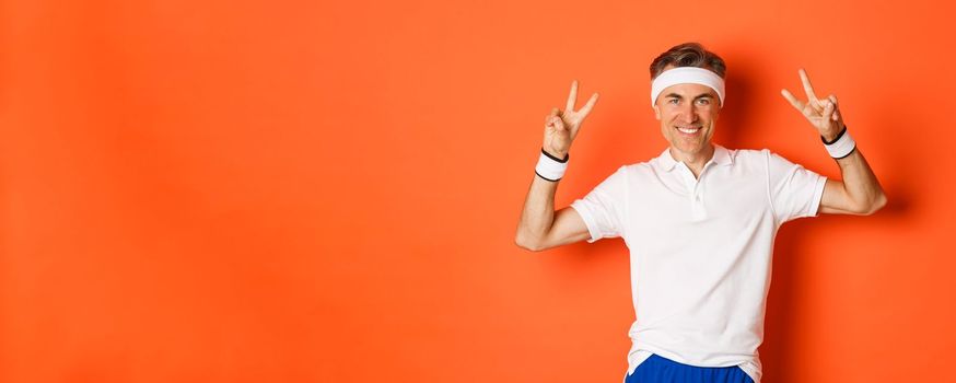 Portrait of handsome, healthy middle-aged male athlete in sportswear, showing peace signs and smiling, workout in gym, standing over orange background