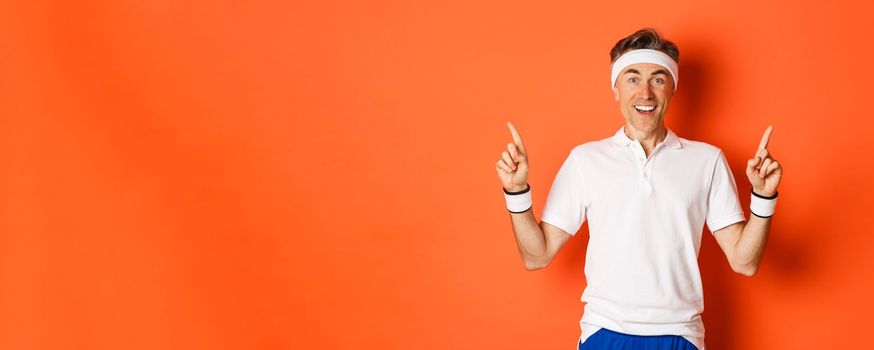 Portrait of healthy, handsome middle-aged male athlete, doing sports and feeling happy, pointing fingers up at logo, standing in activewear over orange background