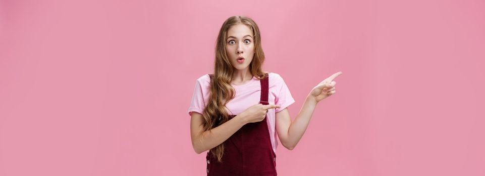 Excited stunned and surprised charming young girl with makeup and curvy hairstyle in trendy corduroy dungarees pointing left folding lips from interest and amazement being curious what happened