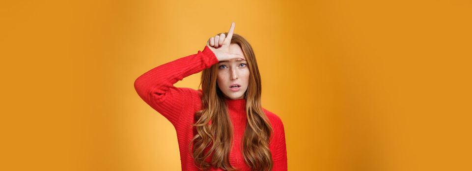 Arrogant and self-satisfied redhead woman humiliating person showing loser sign on forehead mocking and disdain rival standing confident and snobbish over orange background
