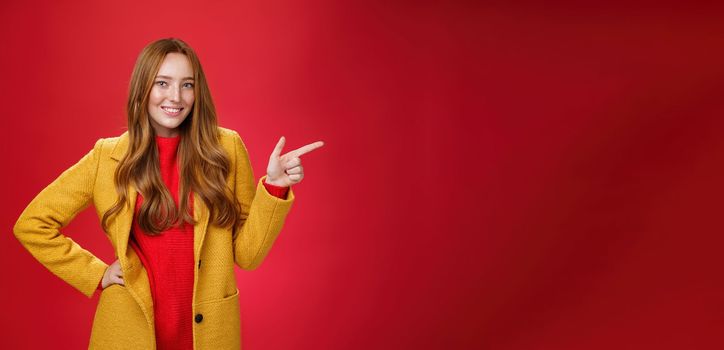 Stylish and self-assured attractive redhead woman with freckles in yellow outdoor coat pointing right with finger pistol and smiling joyfully as camera as showing cool promotion over red wall